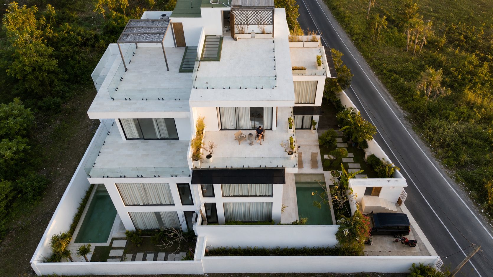 The villa from directly above in golden light — white rooftops and terraces stepping down to the plunge pools, the garden and resort road around it.