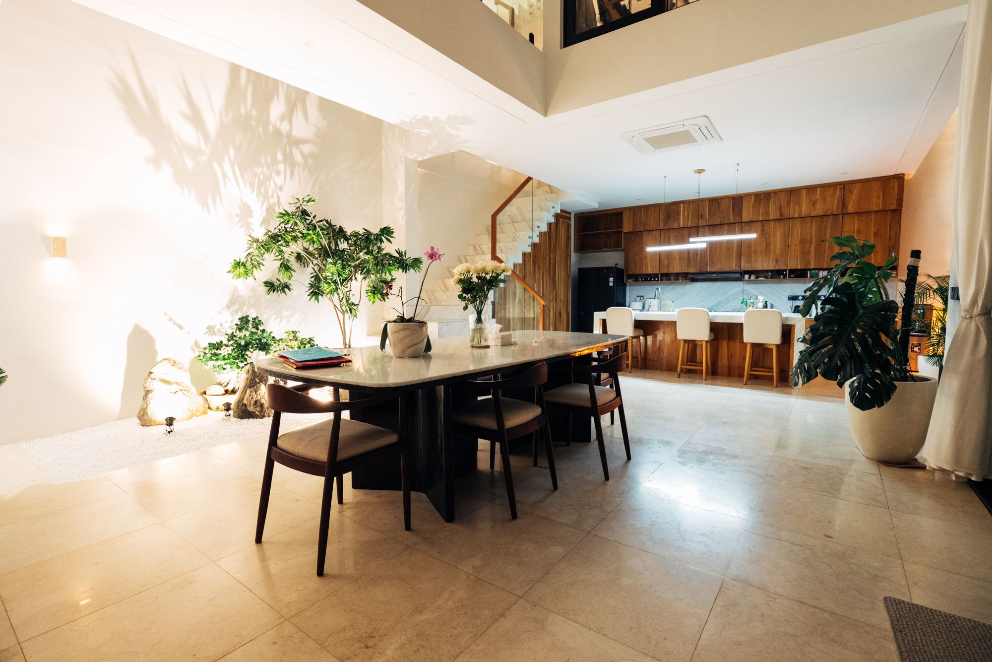 A planted indoor garden lit by a skylight from the roof, beside the marble dining table, the timber kitchen beyond.