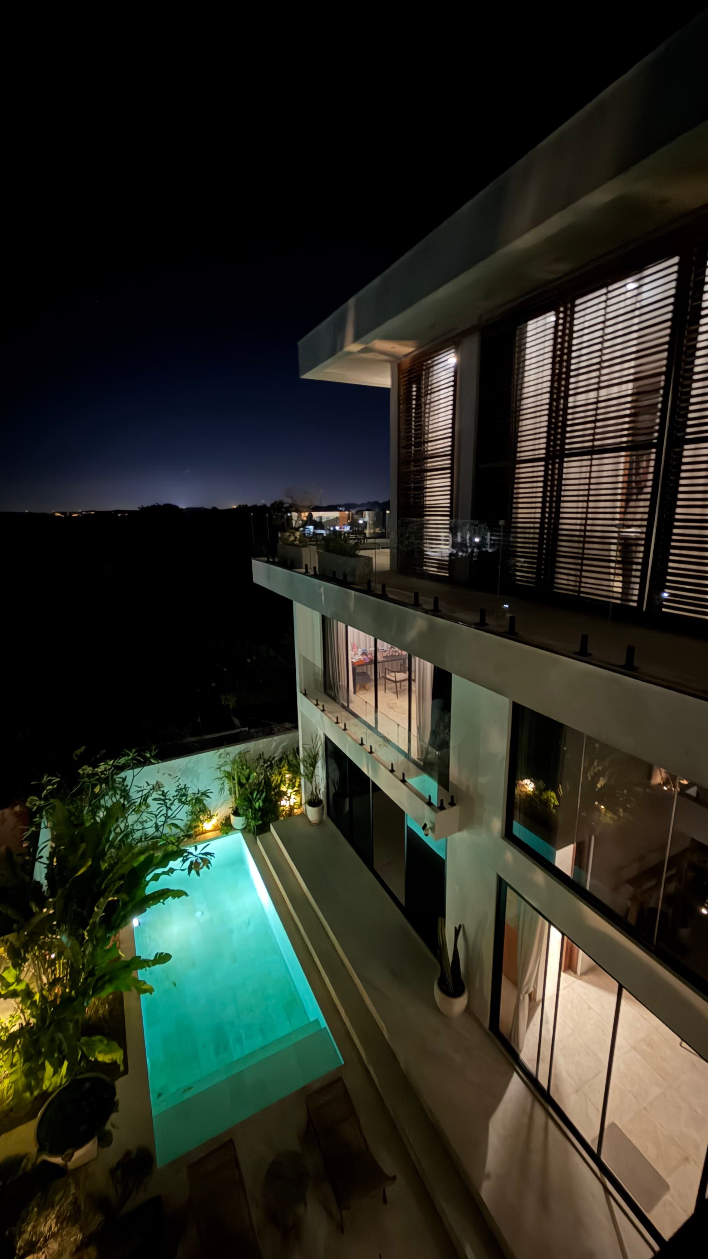 Looking down the side of the lit villa at night: the plunge pool glowing turquoise, warm rooms behind glass, distant coast lights.