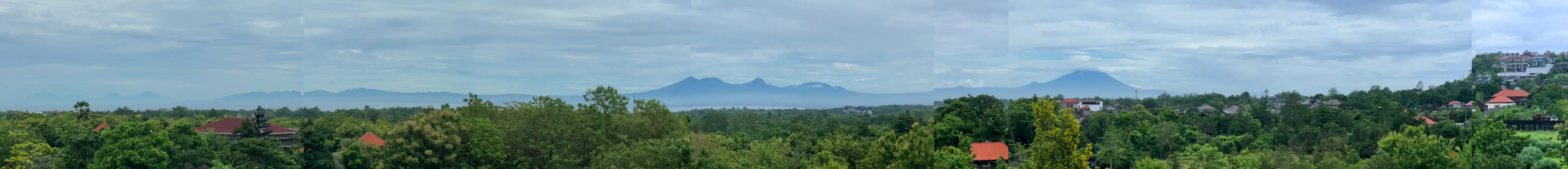 The full terrace outlook in one sweep: the green Bukit and resort rooftops, a band of sea, and the chain of mainland volcanoes with Gunung Agung on the horizon.