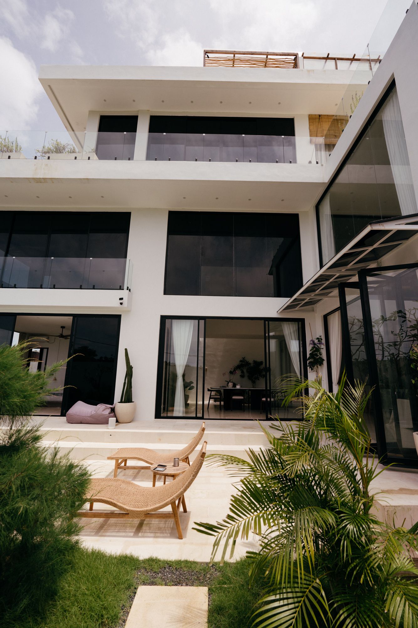 The Sunset villa from the garden — stacked white volumes, black-glass sliders and rattan loungers on the lawn.